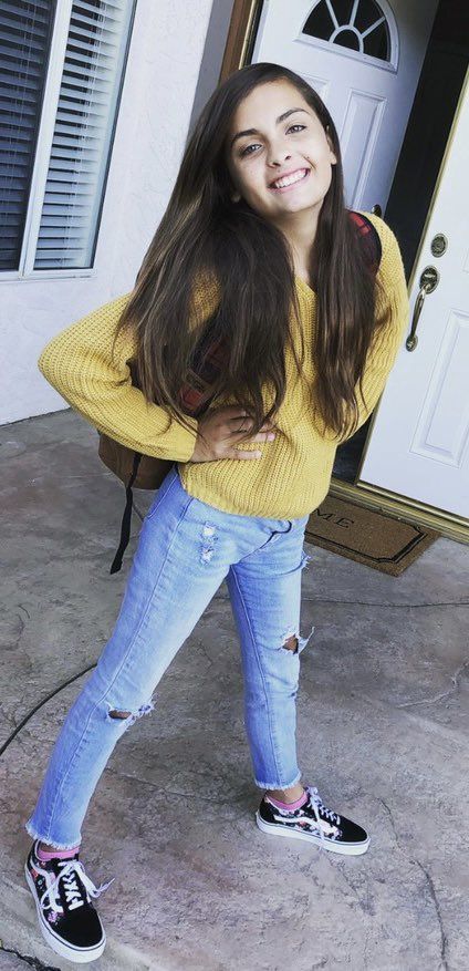 Penny smiling outside her front door on her first day of public school, wearing a yellow top, ripped jeans and black sneakers.